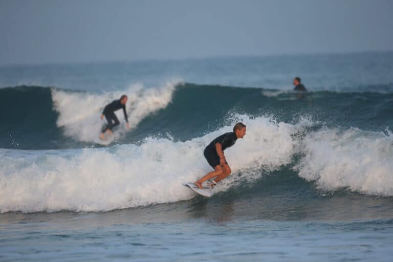 Tre surfere rider på havets bølger. Forgrundssurferen sidder på hug på en bølge midt i bevægelse. En anden surfer er synlig på en bølge i baggrunden, med en tredje i det fjerne. Havet er roligt ud over bølgerne, under en klar himmel.