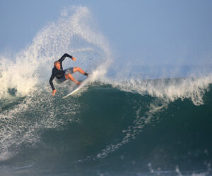 En surfer i en våddragt rider dygtigt på en stor bølge med vand, der sprøjter bag sig. Havet er levende og dynamisk under en klar blå himmel.