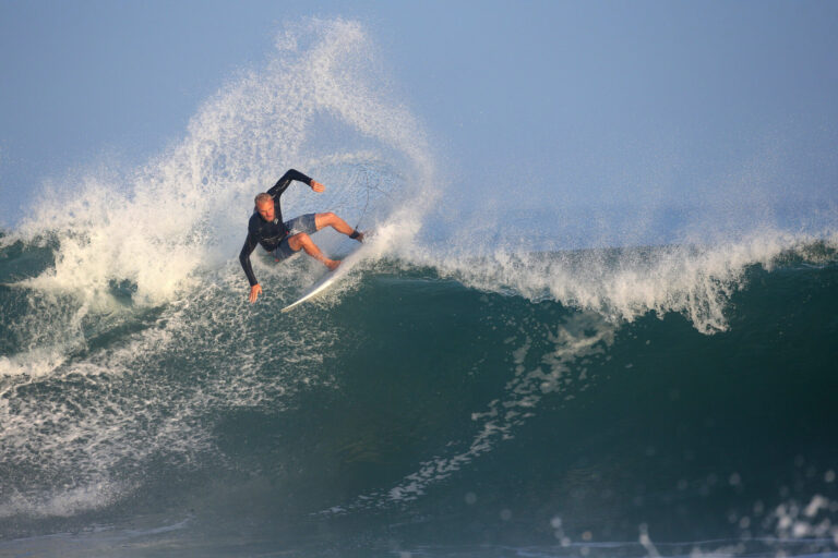 En surfer i en våddragt rider dygtigt på en stor bølge med vand, der sprøjter bag sig. Havet er levende og dynamisk under en klar blå himmel.