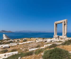 Gamle stenruiner med udsigt over et dybblåt hav under en klar himmel. Marmorporten står fremtrædende på det klippefyldte landskab med fjerne bakker og roligt vand i baggrunden.