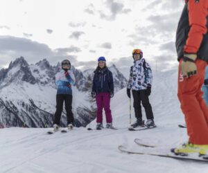 En gruppe skiløbere, iført farverigt skiudstyr, står sammen på en sneklædt bjergskråning. Snedækkede tinder og en overskyet himmel er i baggrunden.