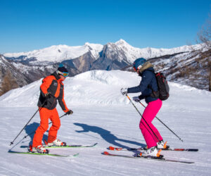 To skiløbere i lyse outfits står på en sneklædt skråning, omgivet af bjerge under en klar blå himmel. Den ene har orange gear, den anden pink. De står over for hinanden og holder skistave med naturskønne tinder i baggrunden.