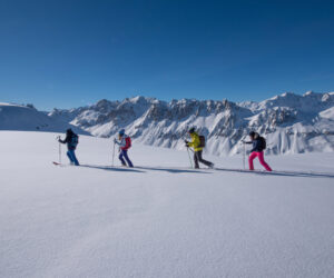 Fire personer på ski op ad bakke på en snedækket bjergskråning under en klar blå himmel. Majestætiske snedækkede tinder er synlige i baggrunden. Skiløberne er iført farverigt vinterudstyr, inklusive jakker og rygsække, der fremhæver en solrig vinterdag.