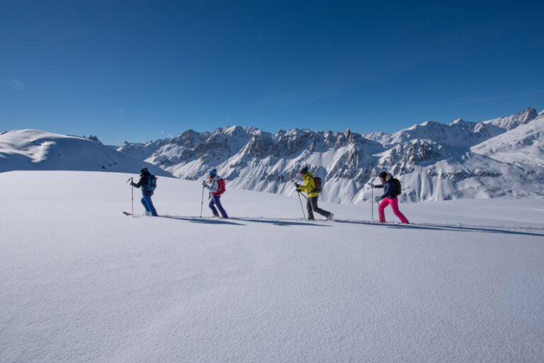 Fire personer på ski op ad bakke på en snedækket bjergskråning under en klar blå himmel. Majestætiske snedækkede tinder er synlige i baggrunden. Skiløberne er iført farverigt vinterudstyr, inklusive jakker og rygsække, der fremhæver en solrig vinterdag.