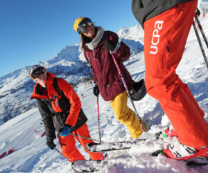 Tre skiløbere i farverigt vinterudstyr står på en sneklædt bjergskråning og smiler og holder stænger. En klar blå himmel og majestætiske snedækkede tinder er synlige i baggrunden. Lyse røde skibukser med UCPA skrevet på dem skiller sig tydeligt ud.