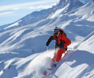 En skiløber i rødt jakkesæt og hjelm går dygtigt ned ad en snedækket bjergskråning og sparker pudder op i luften. Solen skinner klart og oplyser det vidstrakte snelandskab og fjerne tinder under en klar blå himmel.