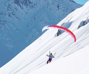 En person paraglider ned ad en snedækket bjergskråning, i kontrast til den klare blå himmel. Den farvestrålende faldskærm er rød og lilla og fanger vintersportens fredfyldte, men alligevel eventyrlystne ånd.