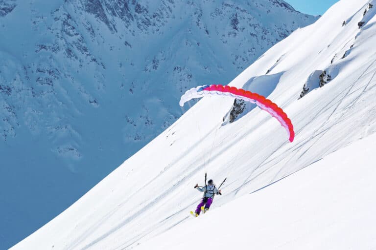En person paraglider ned ad en snedækket bjergskråning, i kontrast til den klare blå himmel. Den farvestrålende faldskærm er rød og lilla og fanger vintersportens fredfyldte, men alligevel eventyrlystne ånd.
