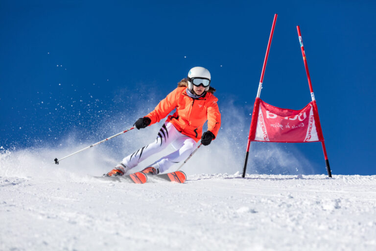 En skiløber i en lys orange jakke og hvide bukser manøvrerer dygtigt rundt om en rød port på en sneklædt skråning under en klar blå himmel. Sne sparkes op bagved, og skiløberen bærer hvid hjelm og beskyttelsesbriller.