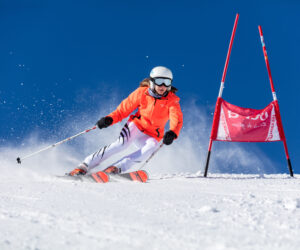 En skiløber i orange jakke og hjelm navigerer på en slalombane på en snedækket skråning med blå himmel i baggrunden. Skiløberen bliver fanget midt i svinget nær en rød portmarkør, hvilket skaber en spray af sne.