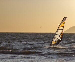 En windsurfer glider over havets bølger under solnedgang med en bjergrig kystlinje i baggrunden. Himlen er en varm gylden nuance, og et par kitesurfere kan ses i det fjerne.