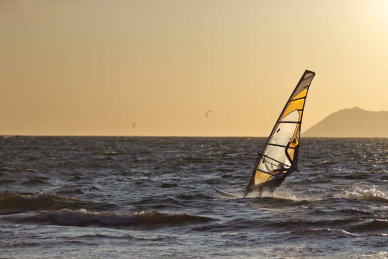 En windsurfer glider over havets bølger under solnedgang med en bjergrig kystlinje i baggrunden. Himlen er en varm gylden nuance, og et par kitesurfere kan ses i det fjerne.