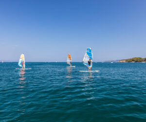 Tre personer, der windsurfer på et roligt, blåt hav under en klar himmel. De er adskilt og balancerer hver især på deres brædder med farverige sejl. Den fjerne kyst er synlig med grønne områder og enkelte spredte både.