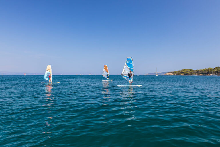 Tre personer, der windsurfer på et roligt, blåt hav under en klar himmel. De er adskilt og balancerer hver især på deres brædder med farverige sejl. Den fjerne kyst er synlig med grønne områder og enkelte spredte både.