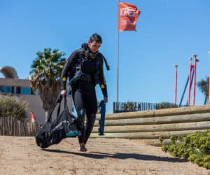 En person i sort dykkerdragt går ad en sandet sti med dykkerudstyr. De har en stor sort taske og et par svømmefødder. Et rødt flag blafrer i baggrunden mod en klar blå himmel.