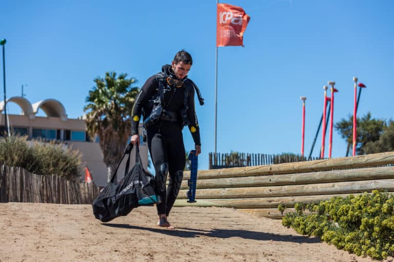 En person i sort dykkerdragt går ad en sandet sti med dykkerudstyr. De har en stor sort taske og et par svømmefødder. Et rødt flag blafrer i baggrunden mod en klar blå himmel.