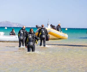 En gruppe dykkere i våddragter og udstyr går gennem lavt vand mod en gummibåd, der er forankret nær kysten. Havet er klart og blåt, og andre både er synlige i baggrunden under en solrig himmel.