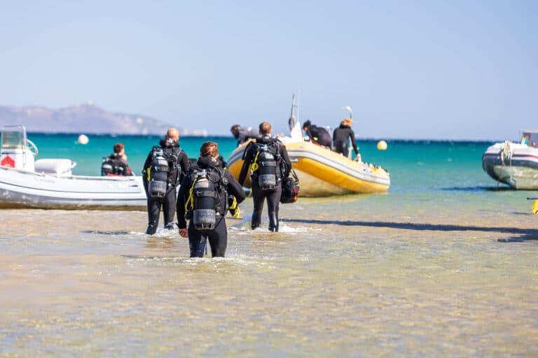 En gruppe dykkere i våddragter og udstyr går gennem lavt vand mod en gummibåd, der er forankret nær kysten. Havet er klart og blåt, og andre både er synlige i baggrunden under en solrig himmel.