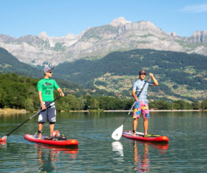 To mennesker padleboarding på en rolig sø med røde brædder, omgivet af frodige grønne bakker og majestætiske bjerge under en klar blå himmel. Den ene har en grøn skjorte og kasket på, den anden i blå skjorte og shorts, begge med solbriller.