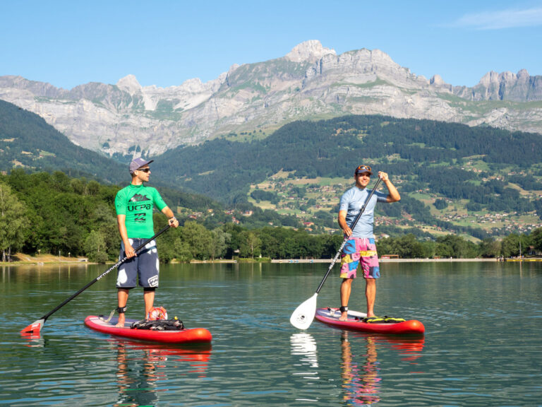 To mennesker padleboarding på en rolig sø med røde brædder, omgivet af frodige grønne bakker og majestætiske bjerge under en klar blå himmel. Den ene har en grøn skjorte og kasket på, den anden i blå skjorte og shorts, begge med solbriller.
