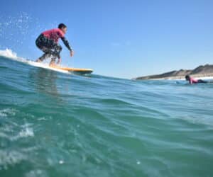 En surfer iført en rød og sort våddragt rider på en bølge på et klart blåt hav. En anden person i baggrunden svømmer på et surfbræt. Himlen er klar, og kystlinjen med sandede bakker er synlig.