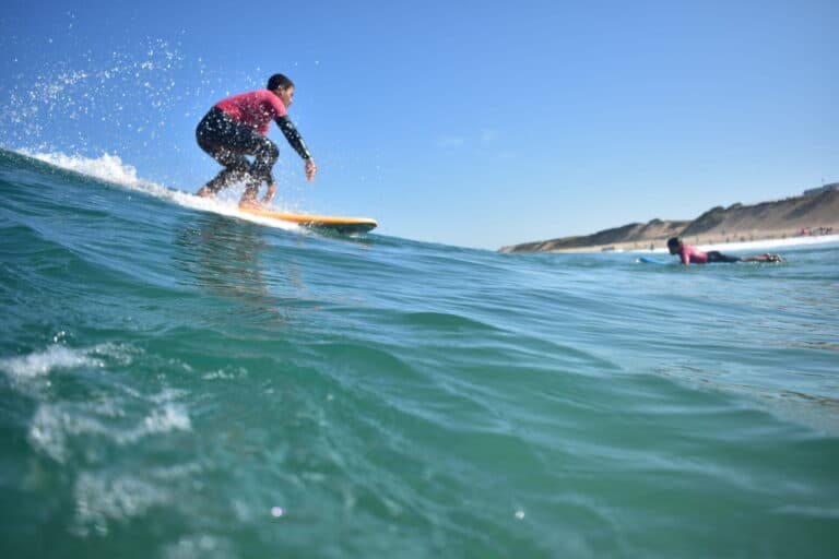 En surfer iført en rød og sort våddragt rider på en bølge på et klart blåt hav. En anden person i baggrunden svømmer på et surfbræt. Himlen er klar, og kystlinjen med sandede bakker er synlig.