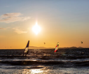 Windsurfere glider hen over havet under en gylden solnedgang. Silhuetter af bjerge langs horisonten, og drager kan ses på himlen, hvilket bidrager til den naturskønne udsigt. Bølger bølger blidt under solens varme skær.
