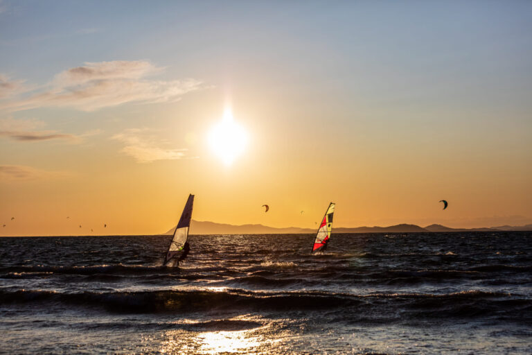 Windsurfere glider hen over havet under en gylden solnedgang. Silhuetter af bjerge langs horisonten, og drager kan ses på himlen, hvilket bidrager til den naturskønne udsigt. Bølger bølger blidt under solens varme skær.