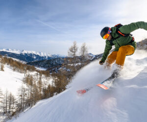 En skiløber i en grøn jakke og gule bukser går ned ad en sneklædt bjergskråning og skaber en spray af sne. Baggrunden byder på snedækkede bjerge og træer under en klar blå himmel.