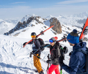 Tre mennesker i skiudstyr går langs en sneklædt bjergryg, bærende ski over skuldrene og nyder deres skiferie. De smiler og sludrer under en klar blå himmel med panoramaudsigt over snedækkede tinder i baggrunden.