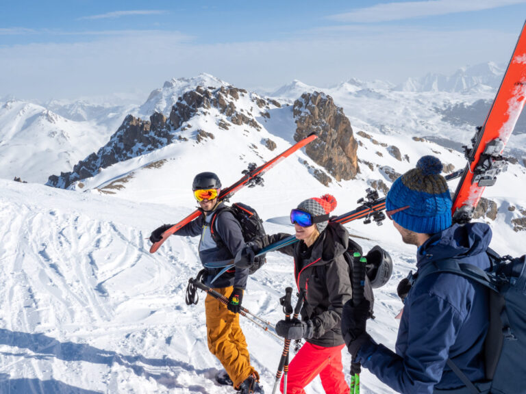 Tre mennesker i skiudstyr går langs en sneklædt bjergryg, bærende ski over skuldrene og nyder deres skiferie. De smiler og sludrer under en klar blå himmel med panoramaudsigt over snedækkede tinder i baggrunden.