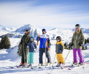 En gruppe på fem personer i skiudstyr står på en sneklædt skråning med ski og stænger. Snedækkede bjerge og en klar blå himmel er i baggrunden. Træer præger landskabet.