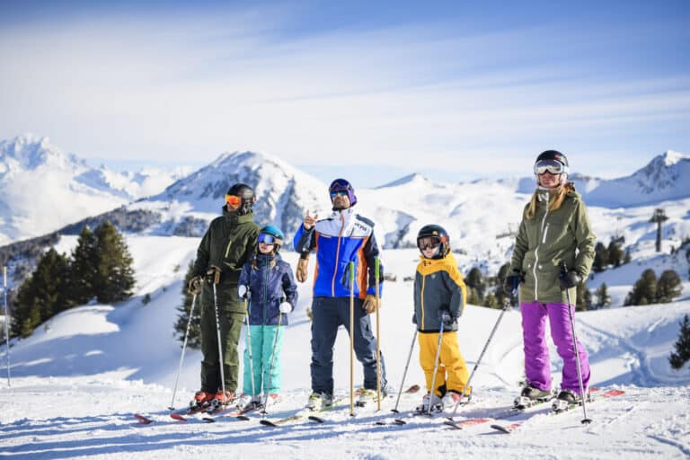 En gruppe på fem personer i skiudstyr står på en sneklædt skråning med ski og stænger. Snedækkede bjerge og en klar blå himmel er i baggrunden. Træer præger landskabet.