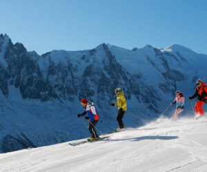Fire skiløbere går ned ad en sneklædt bjergskråning. De er klædt i farverige jakker på baggrund af majestætiske, barske tinder under en klar blå himmel. Solen fremhæver den friske puddersne og skaber en malerisk vinterscene.