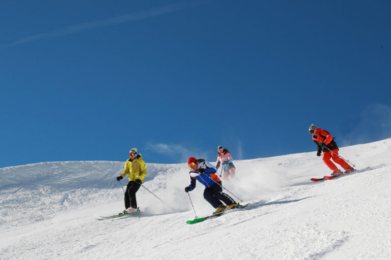 Fire skiløbere i farverige outfits glider ned ad en sneklædt skråning under en klar blå himmel. De er arrangeret i en forskudt formation, der hver viser forskellige skistile og skaber spor af sne bag dem.
