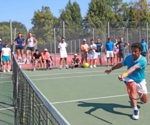 En tennisspiller i blåt og orange slår en forehand nær nettet, mens en gruppe mennesker med ketsjere ser til fra sidelinjen. Scenen foregår på en solskinsdag på en udendørs tennisbane.