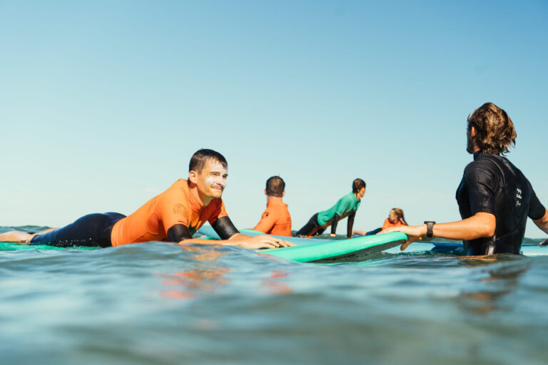 En gruppe mennesker iført orange og grønne udslætsvagter svæver på surfbrætter i havet. De er engageret i en surflektion under en klar blå himmel, hvor en person instruerer de andre.