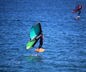 To personer på hydrofoilbrædder manøvrerer hen over havet. Den tættere person holder en grøn og sort vinge, mens den fjernere har en orange og sort vinge. Begge er iført våddragter og glider hen over det dybblå vand.
