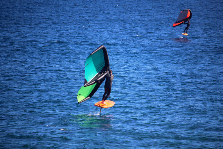 To personer på hydrofoilbrædder manøvrerer hen over havet. Den tættere person holder en grøn og sort vinge, mens den fjernere har en orange og sort vinge. Begge er iført våddragter og glider hen over det dybblå vand.