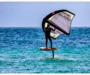 En person, der er engageret i vingefoliering, glider over havets bølger. De står på et bræt fastgjort til en hydrofoil og holder en vinge i vejret. Vandet er klart blåt, og scenen formidler en følelse af frihed og eventyr.