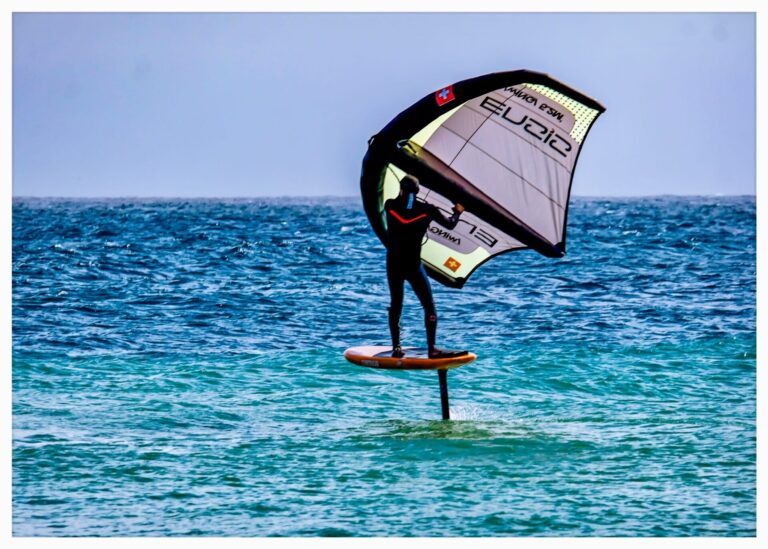 En person, der er engageret i vingefoliering, glider over havets bølger. De står på et bræt fastgjort til en hydrofoil og holder en vinge i vejret. Vandet er klart blåt, og scenen formidler en følelse af frihed og eventyr.