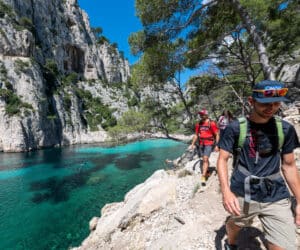 To personer vandrer på en stenet sti ved siden af et klart turkisblåt vand. De er omgivet af stejle klipper og frodige grønne områder under en klar blå himmel. Begge er iført afslappet vandreudstyr og solbriller.
