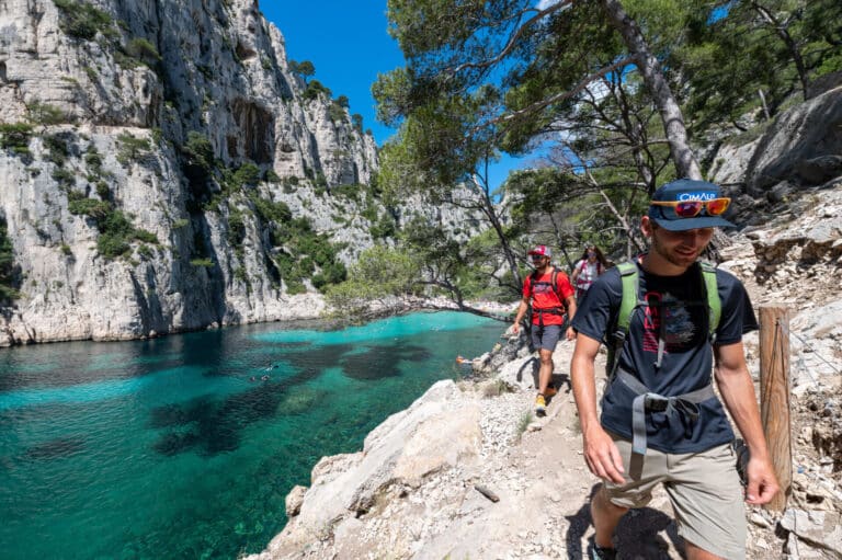 To personer vandrer på en stenet sti ved siden af et klart turkisblåt vand. De er omgivet af stejle klipper og frodige grønne områder under en klar blå himmel. Begge er iført afslappet vandreudstyr og solbriller.