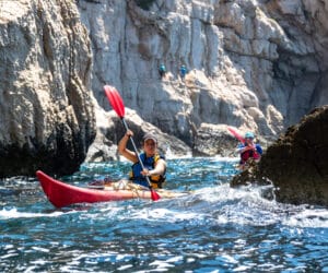 To personer, der sejler i kajak i klart blåt vand nær klippeklipper. Den forreste kajakroer bærer hjelm og padler en rød kajak, mens en anden følger tæt efter. Sollys oplyser scenen og fremhæver det barske landskab og livlige bølger.
