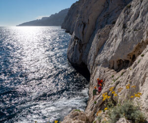 En person i klatreudstyr bestiger en klippeklippe ved siden af et glitrende blåt hav under en klar himmel. Gule blomster vokser på klipperne, og sollyset kaster refleksioner på vandet.