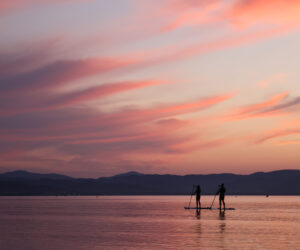 To personer padleboarding på en rolig vandmasse under solnedgang. Himlen er fyldt med lyserøde og orange nuancer, og fjerne bjerge er synlige i horisonten, hvilket skaber en fredfyldt og malerisk scene.