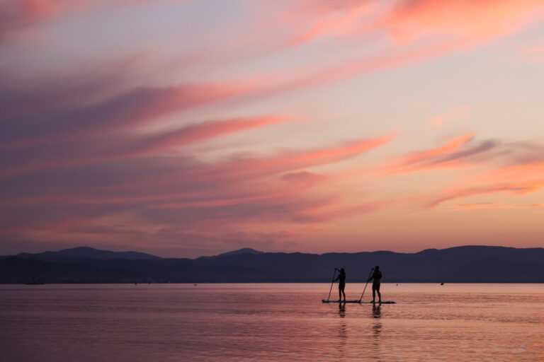 To personer padleboarding på en rolig vandmasse under solnedgang. Himlen er fyldt med lyserøde og orange nuancer, og fjerne bjerge er synlige i horisonten, hvilket skaber en fredfyldt og malerisk scene.