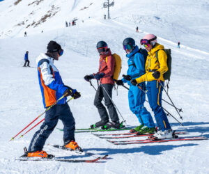 Fire skiløbere i farverige outfits står sammen på en sneklædt skråning med en baggrund af snedækkede bakker og klarblå himmel. De ser ud til at være i samtale og holde skistave.