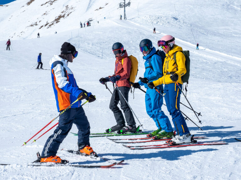 Fire skiløbere i farverige outfits står sammen på en sneklædt skråning med en baggrund af snedækkede bakker og klarblå himmel. De ser ud til at være i samtale og holde skistave.