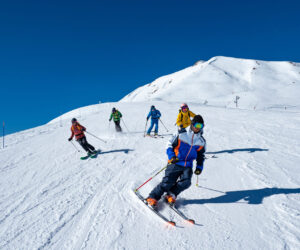 En gruppe skiløbere iført farverige tøj går ned ad en snedækket skråning på en solskinsdag. Himlen er klar, og en sneklædt bjergtop er synlig i baggrunden. De står på ski i en V-formation og skaber en dynamisk actionscene.